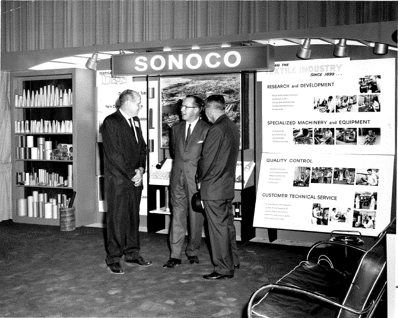 An old black and white photo of three businessmen standing in front of a SONOCO display.