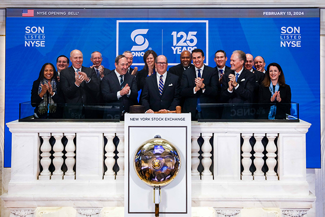 Group of business people wearing suits smiling and clapping in front of a wall that reads, "SON LISTED NYSE, SONOCO 125 Years" Standing in front of the New York Stock Exchange bell.